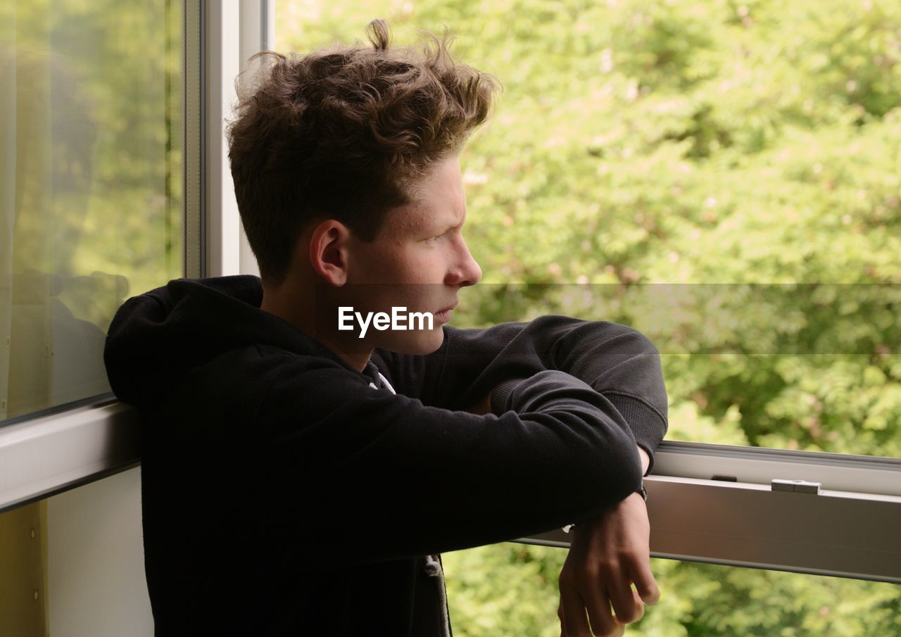 Side view of thoughtful young man looking through window at home