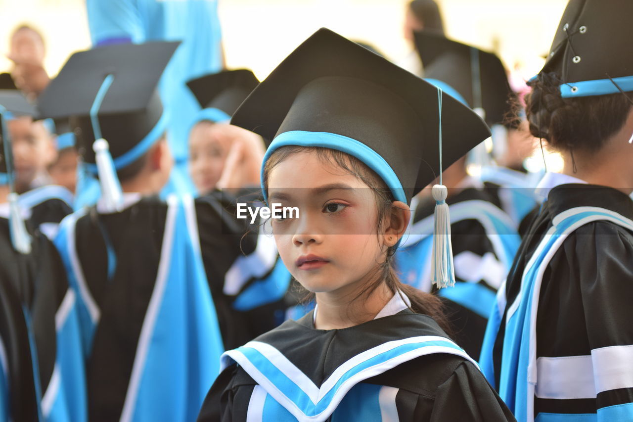 Close-up of girl wearing graduation gown and mortarboard looking away by friends