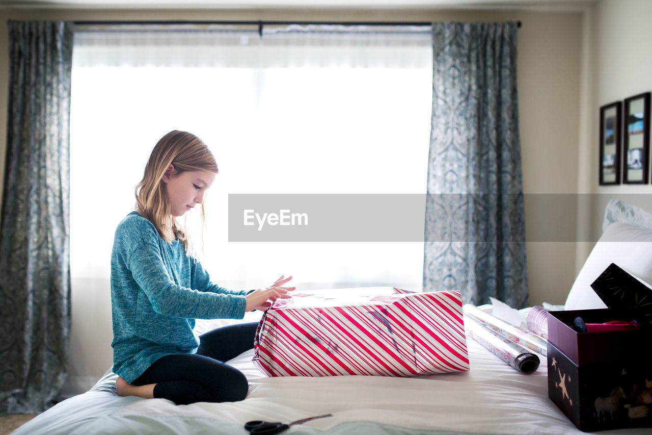 Side view of girl wrapping gift box while kneeling on bed