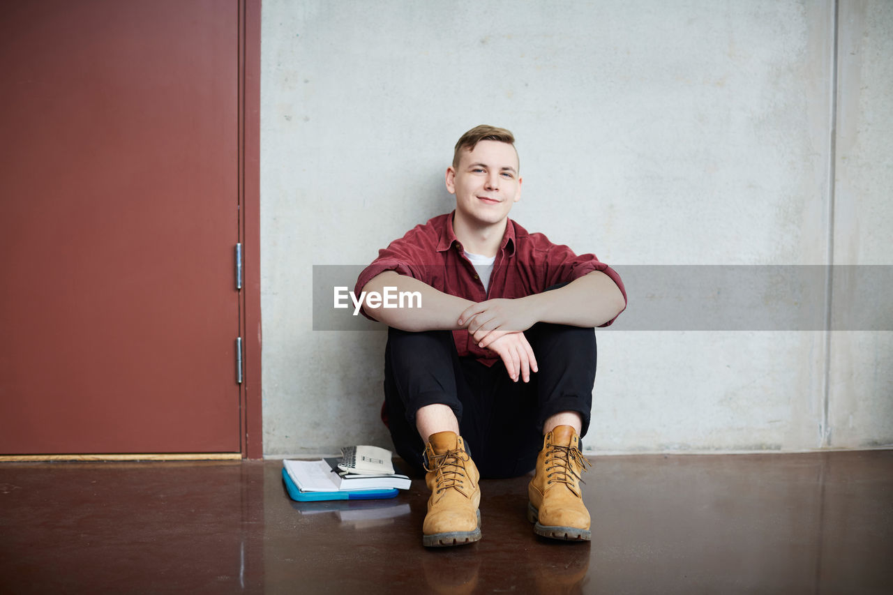 Full length portrait of male student sitting on floor against wall in university