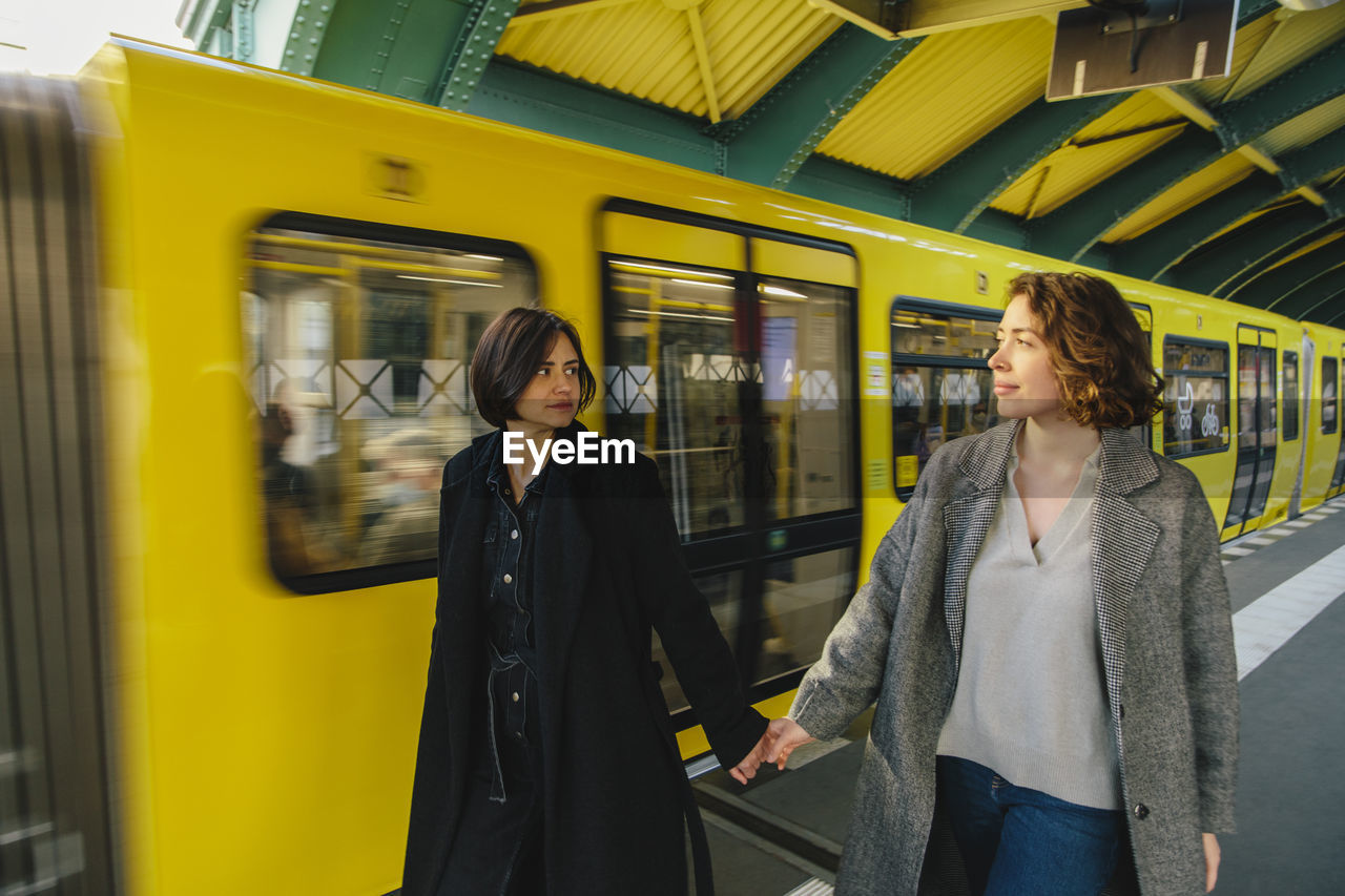 Lesbian couple hand hands walking by yellow train at railroad station