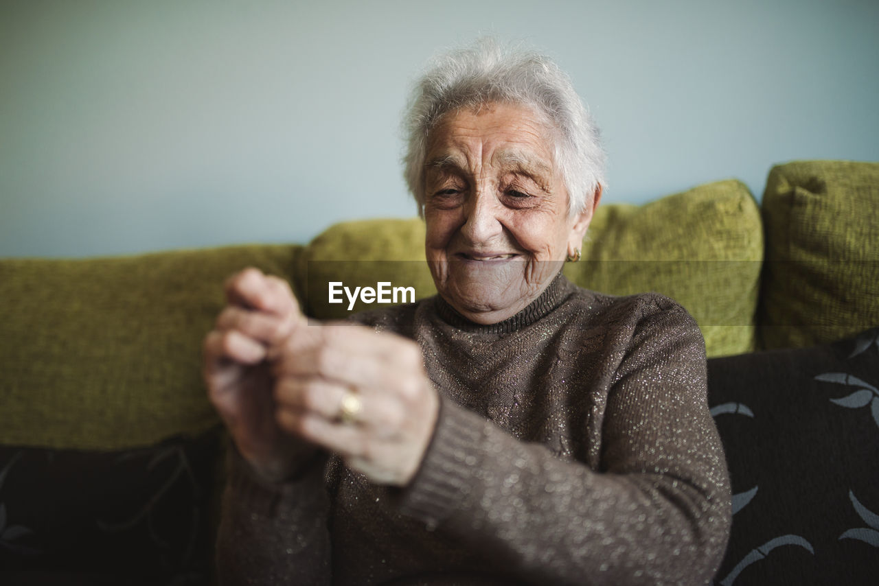 Portrait of smiling senior woman sitting on couch passing thread through buttonhole of sewing needle