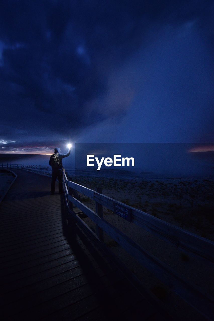 MAN STANDING ON PIER BY SEA AGAINST SKY DURING SUNSET