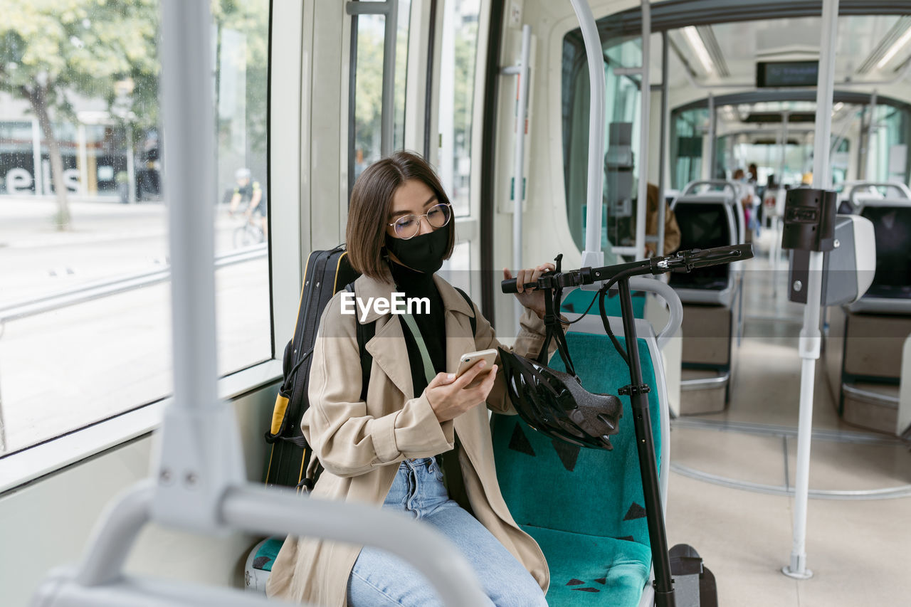 Mid adult woman wearing face mask using mobile phone while sitting with electric push scooter in tram