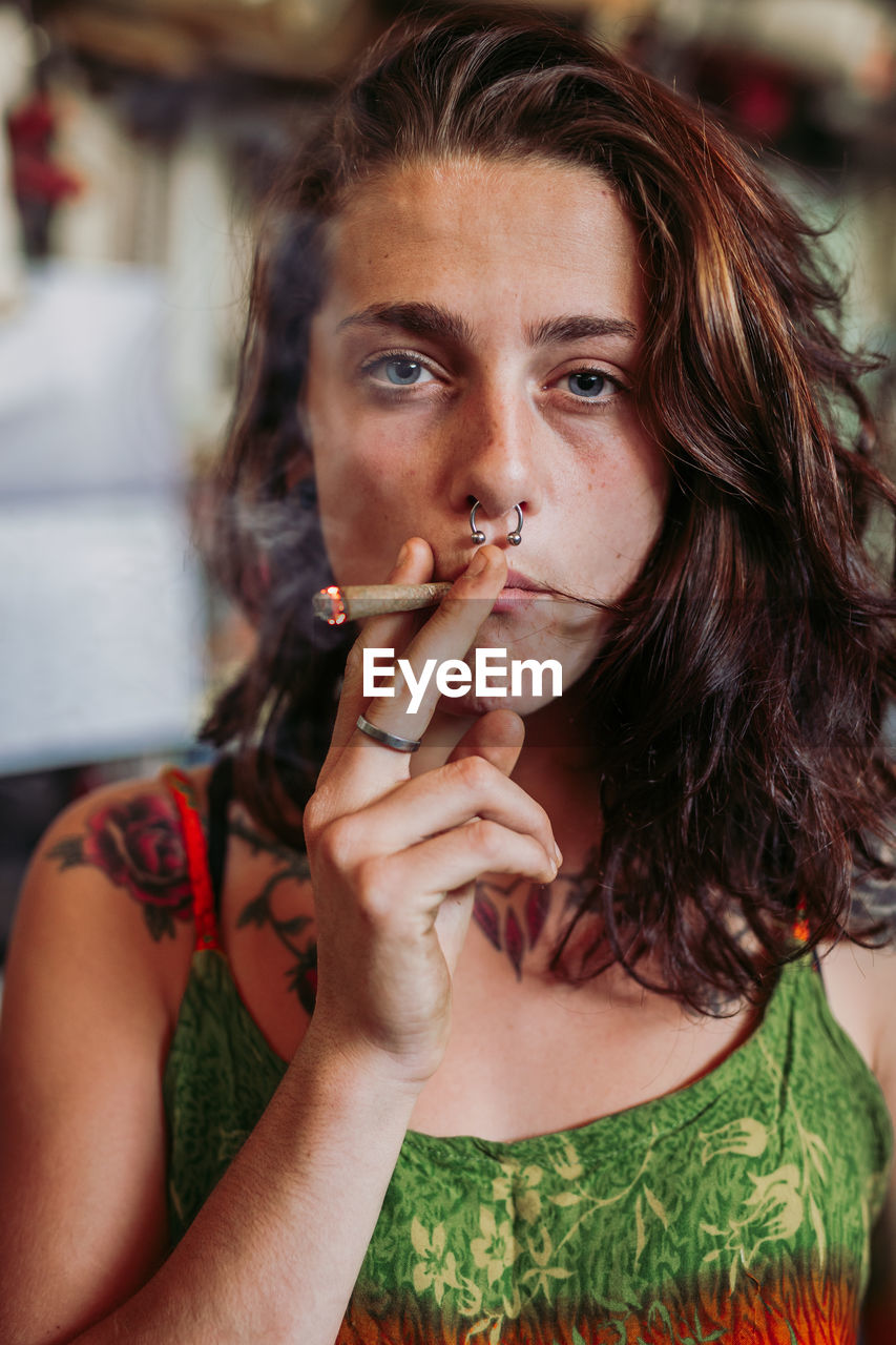 Young red haired female hippie in colorful dress smoking blunt in stylish room looking at camera