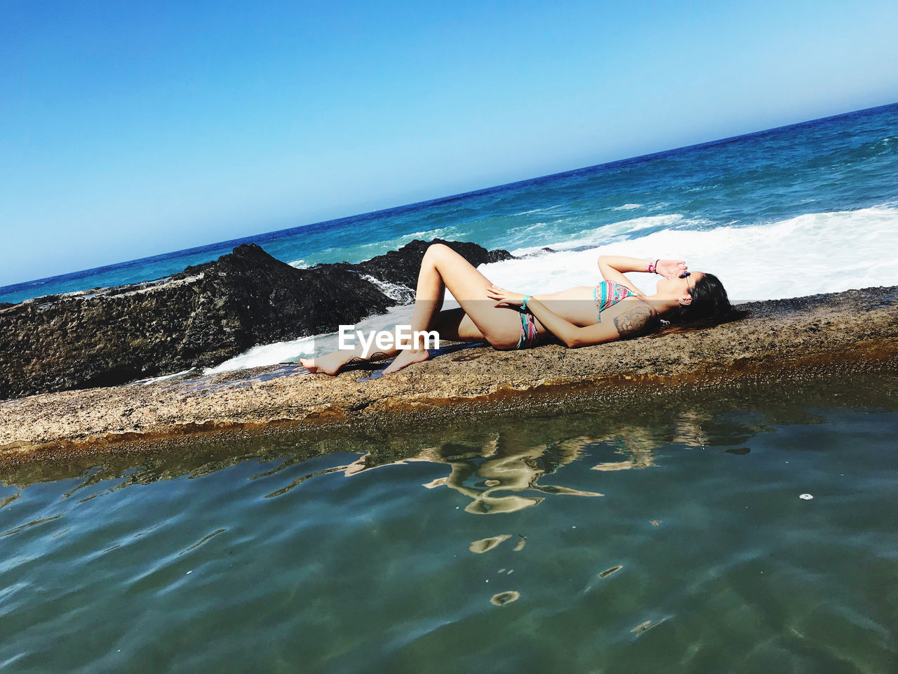 Woman relaxing on retaining wall by sea against clear blue sky