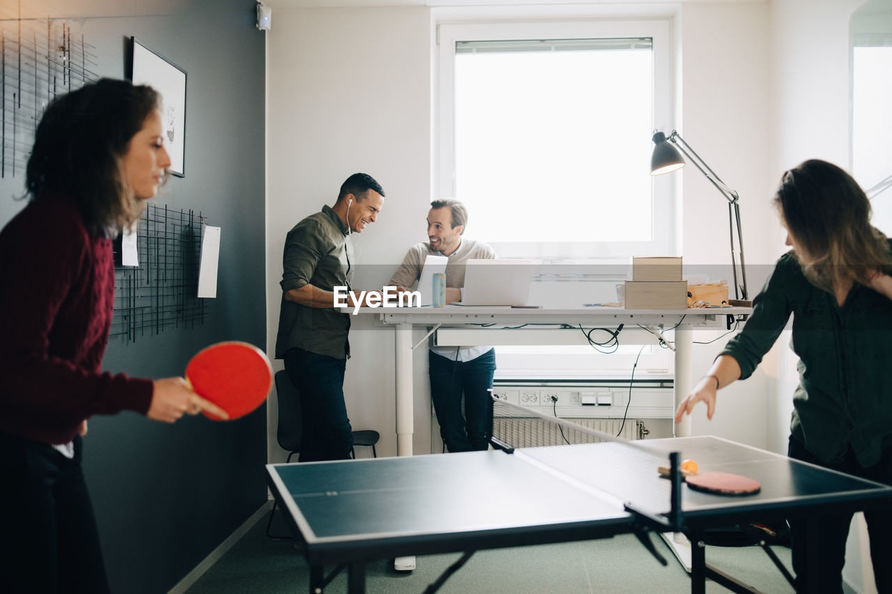 Female professionals playing table tennis while male colleagues using laptop at creative office