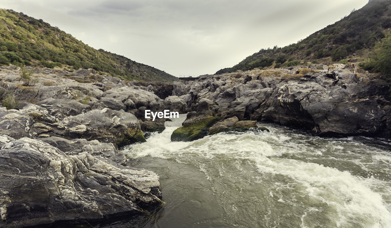 Foam in the rapids of the river with rock covered with green and yellow moss with cloudy sky