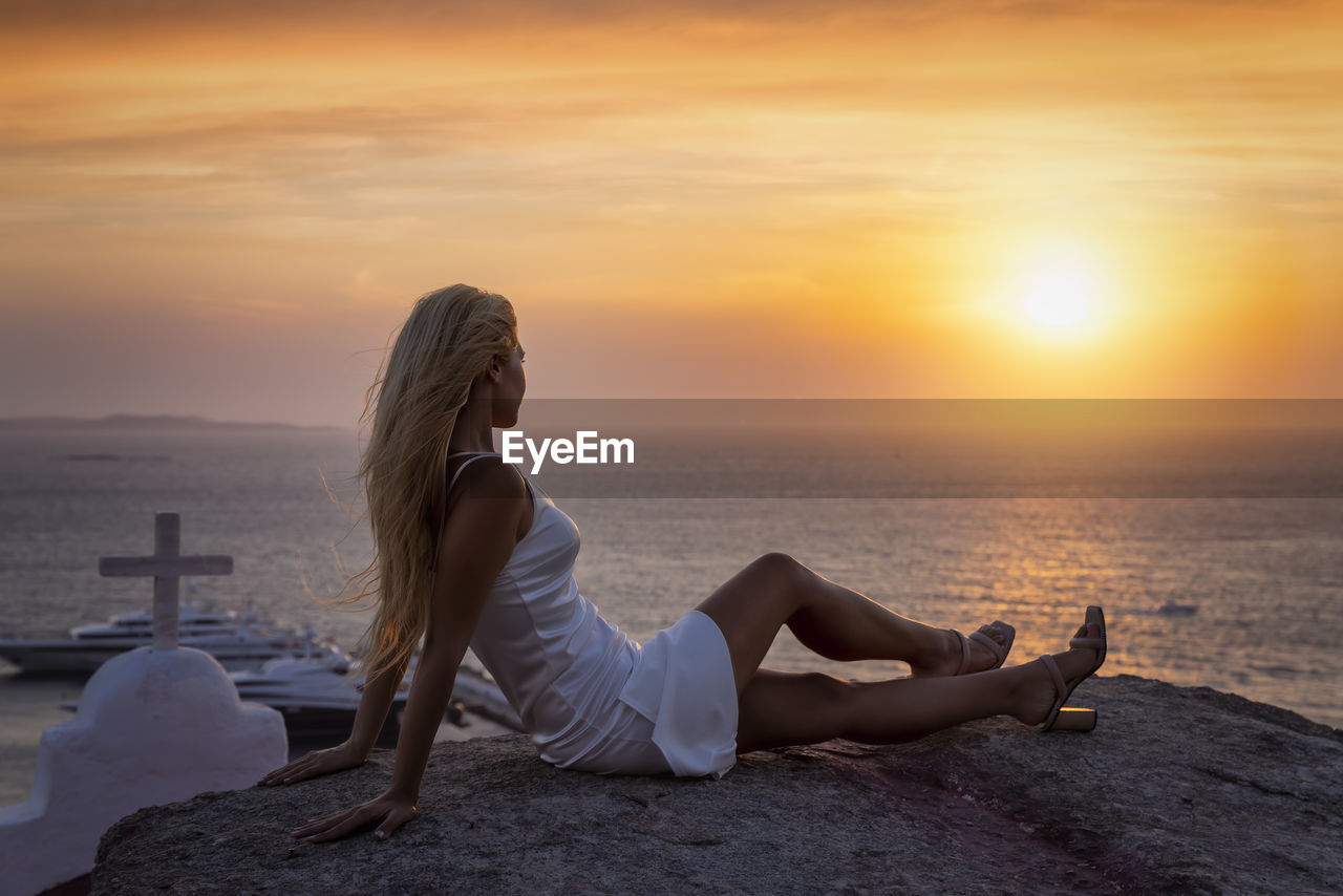 side view of woman sitting on beach against sky during sunset