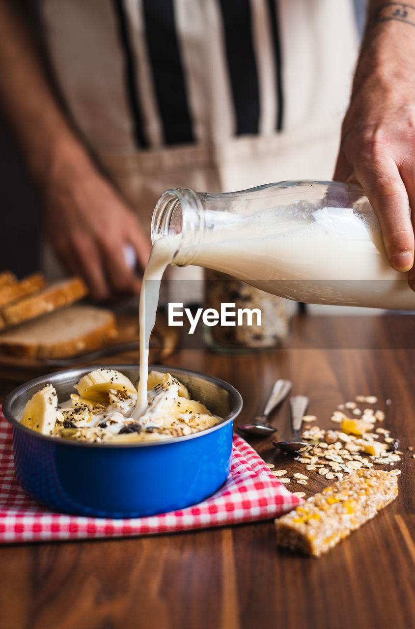 Crop anonymous housewife pouring yogurt from glass bottle into bowl with cereals and banana while preparing healthy breakfast at home kitchen