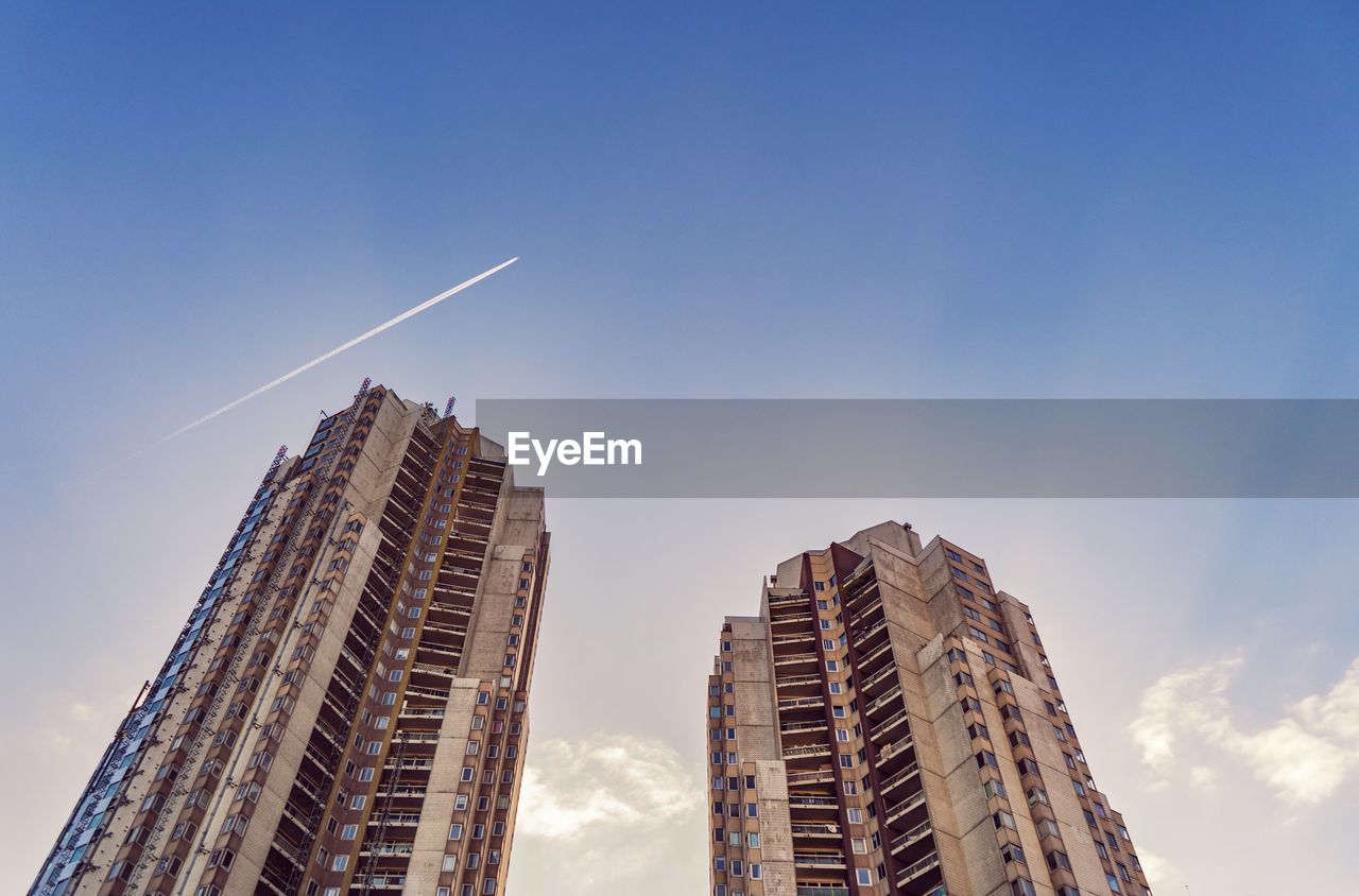 Low angle view of modern buildings against blue sky, plane flying through.