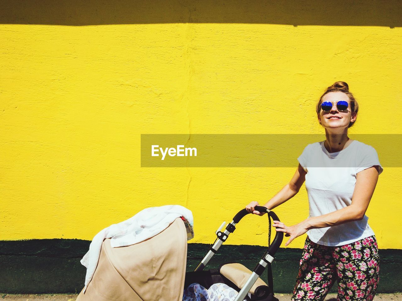 Portrait of woman in sunglasses with baby stroller standing against yellow wall