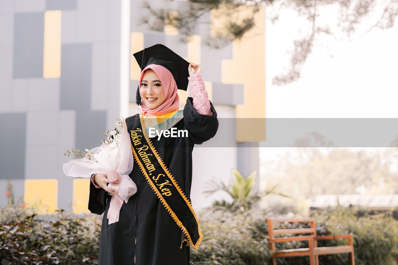 Portrait of young woman wearing graduation gown holding bouquet