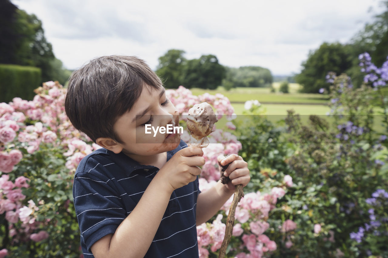 Cute boy eating ice cream sitting against flowering plants