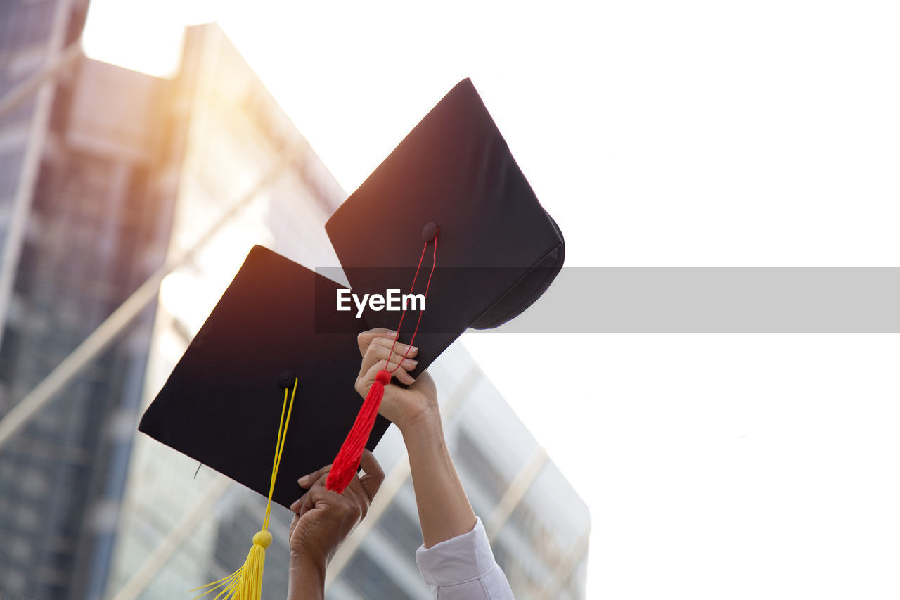 Low angle view of people hands holding mortarboards against sky