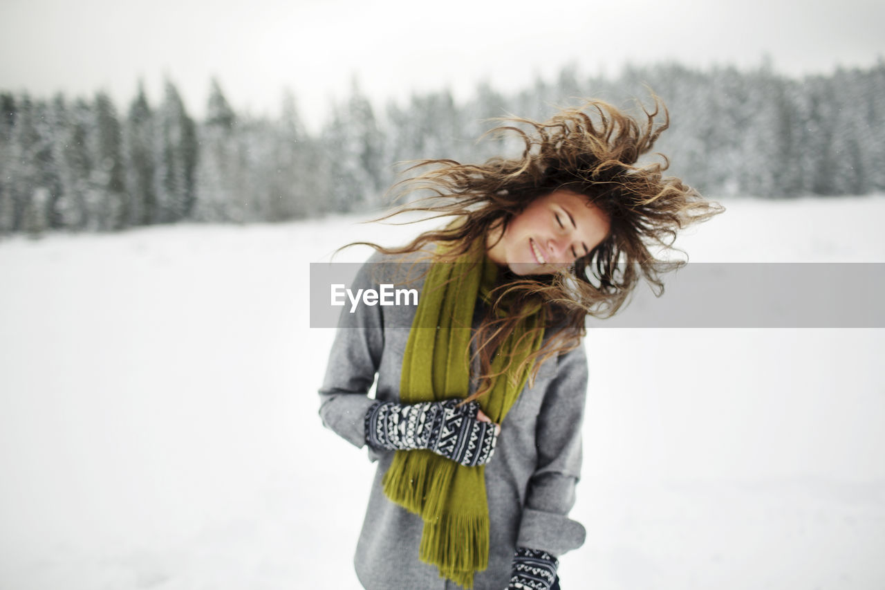 Happy woman tossing hair while standing on snow covered field in forest