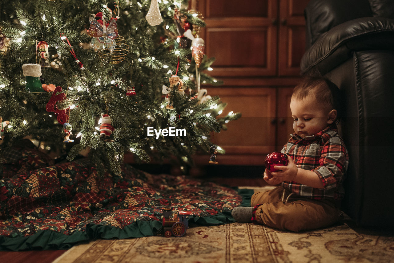 Toddler boy sitting under christmas tree holding ornament