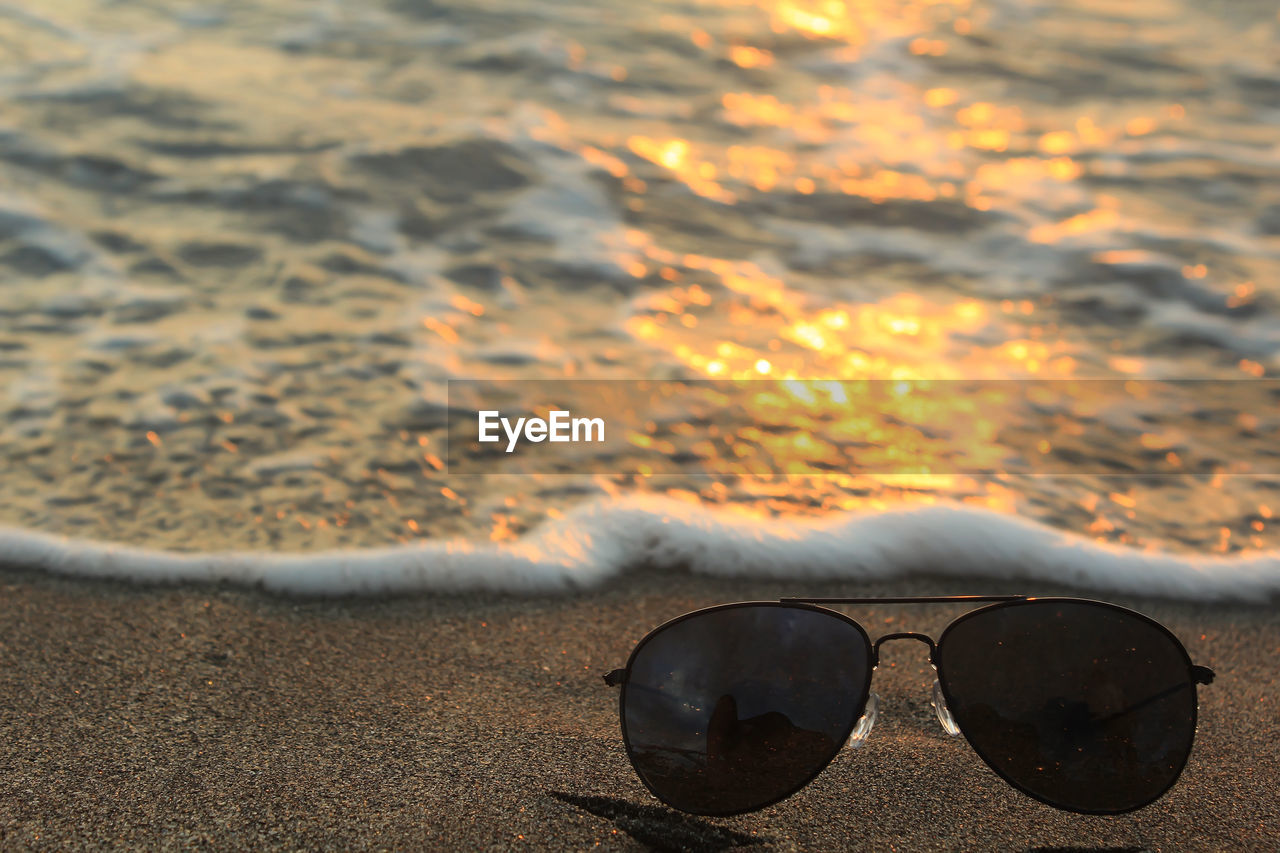CLOSE-UP OF SUNGLASSES ON BEACH AGAINST SKY DURING SUNSET