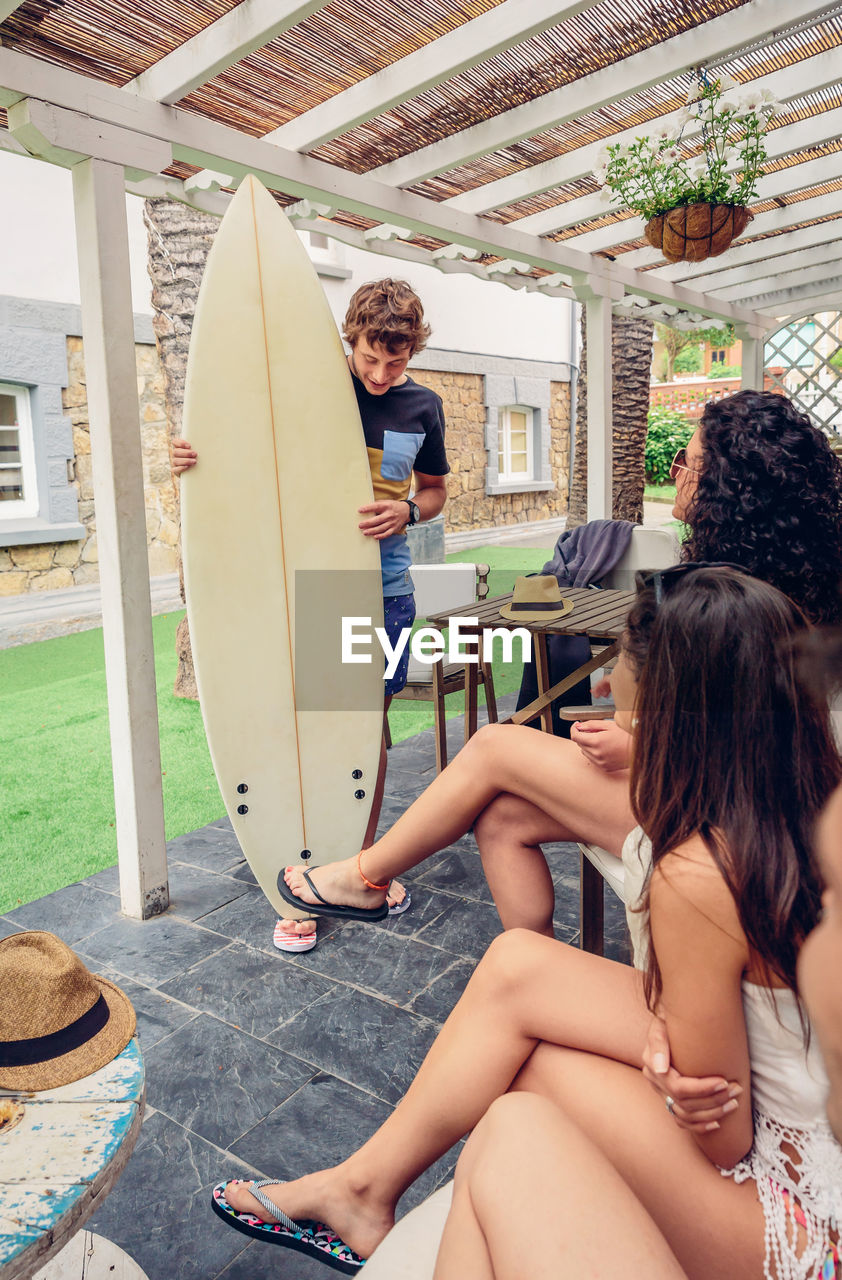 Young female friends looking at man holding surfboard