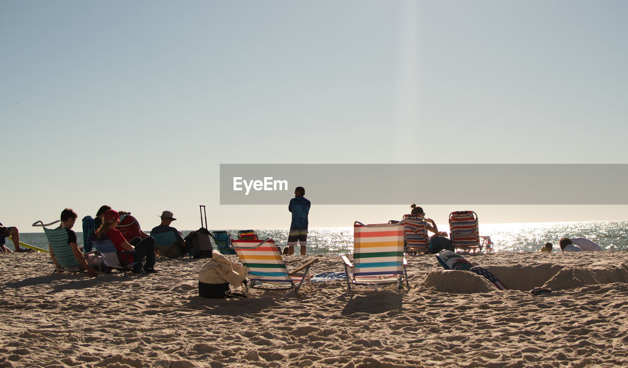 group of people, beach, sky, land, sand, nature, men, adult, copy space, sunlight, women, clear sky, sea, crowd, holiday, trip, vacation, large group of people, leisure activity, sitting, water, summer, day, sunny, relaxation, coast, togetherness, outdoors, lifestyles, group, travel, body of water, standing, natural environment, chair, weekend activities, enjoyment