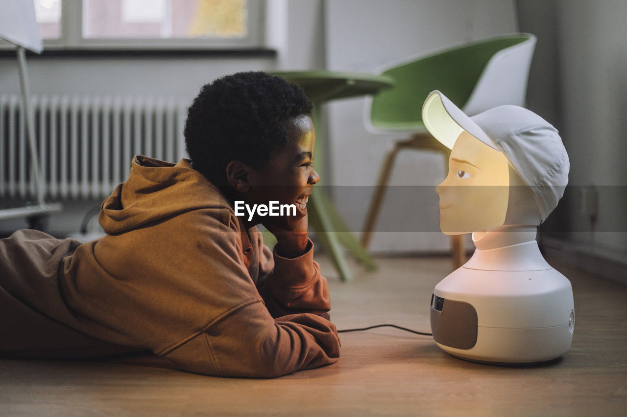 Cheerful boy in front of illuminated ai robot while lying on floor in innovation lab