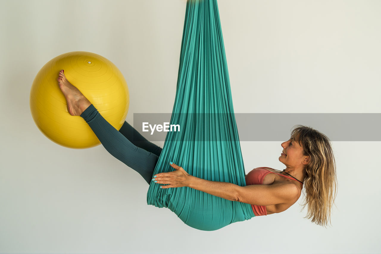 Woman hanging on aerial silk holding fitness ball with legs in front of wall