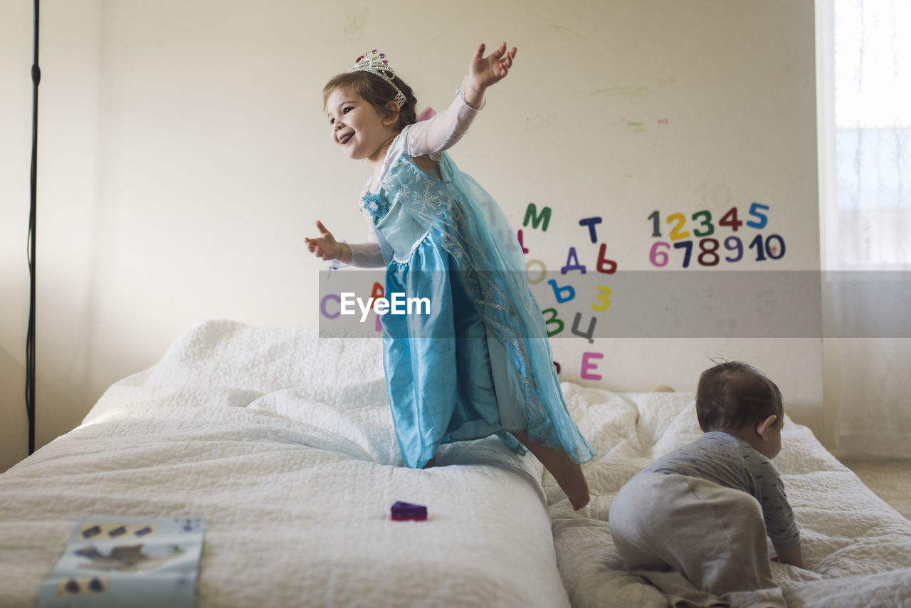 Happy girl wearing princess costume jumping on bed with baby brother negative space
