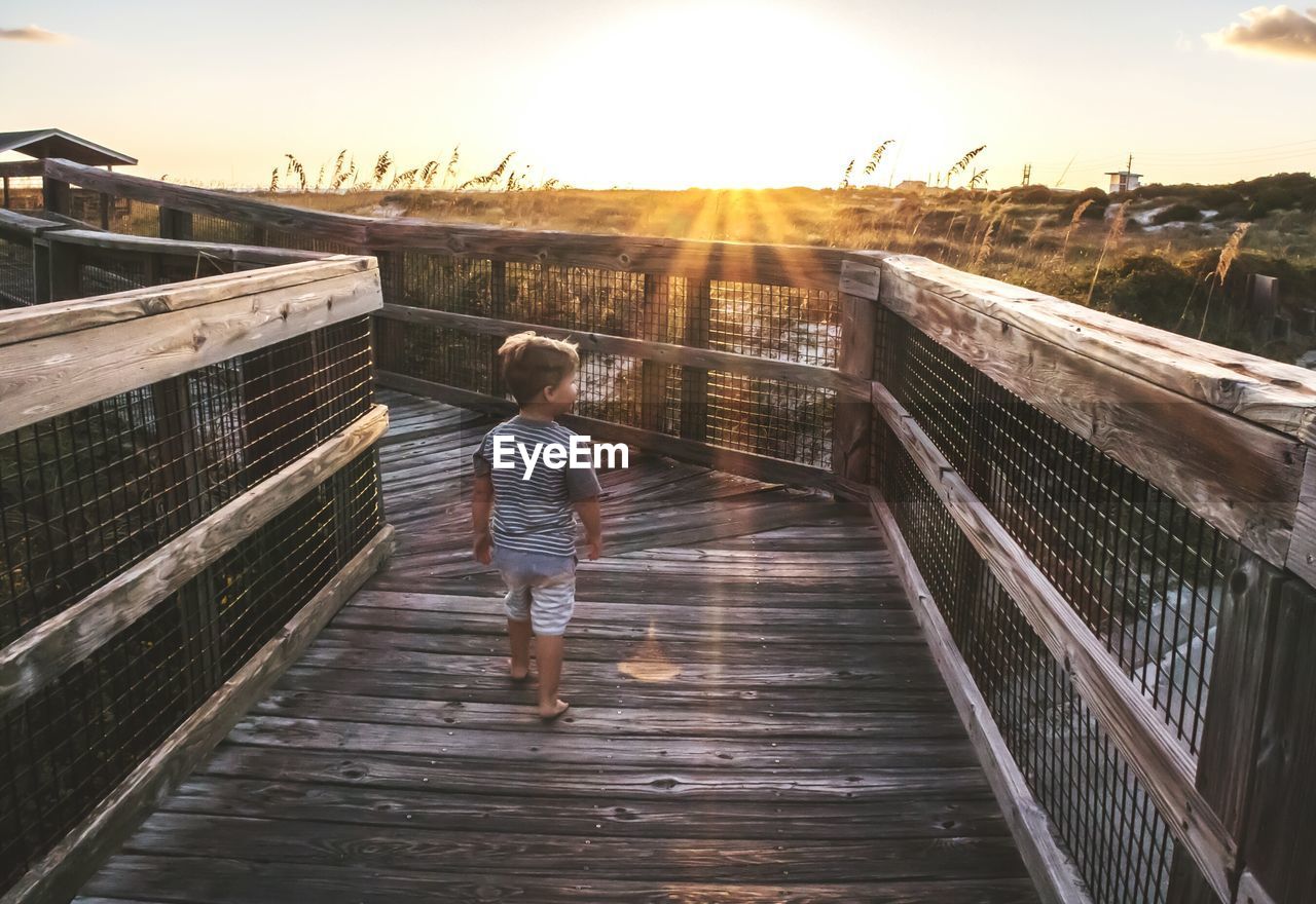 Rear view of little boy walking on boardwalk over field during sunset