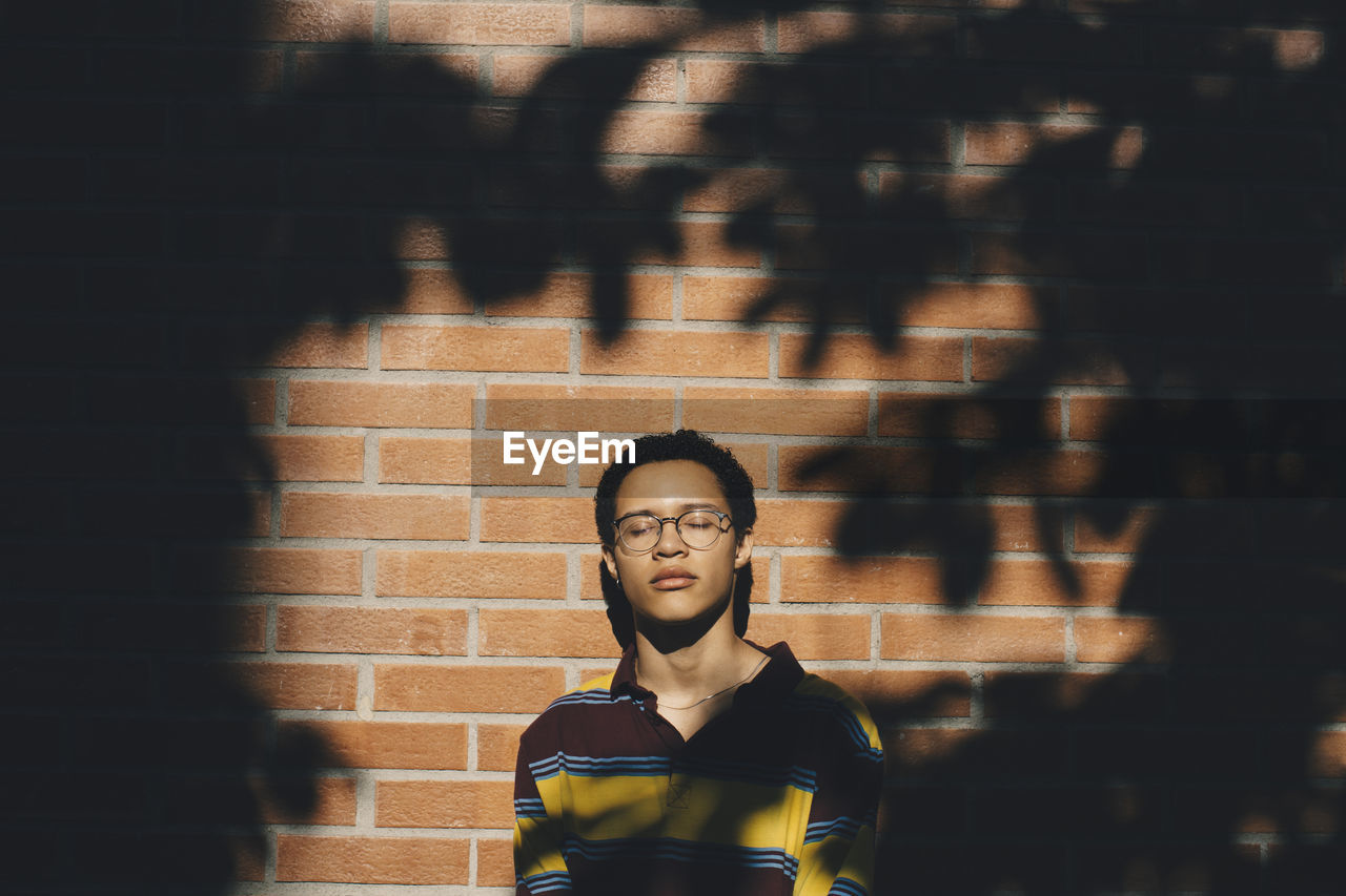 Young man with eyes closed wearing eyeglasses against brick wall