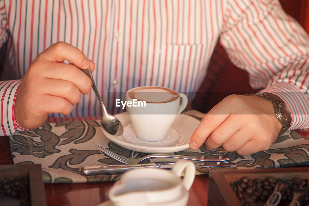 A man in a striped shirt holds a silver teaspoon and a white cup of coffee