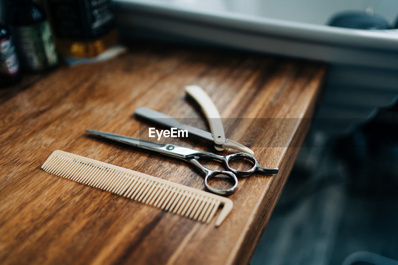 From above scissor and comb near straight razor with sharp metal blades on wooden table in hairdressing salon