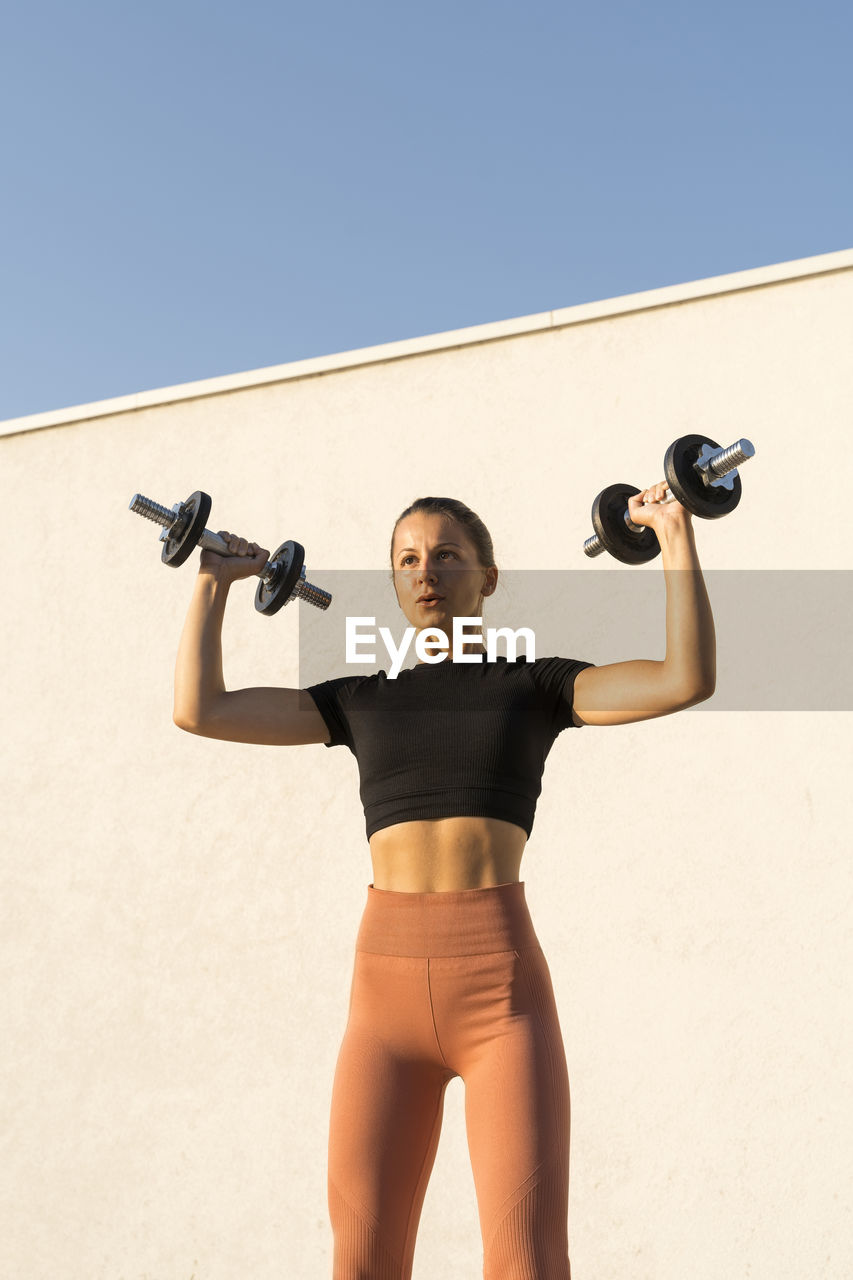 Female athlete flexing muscles with dumbbell standing against wall during sunrise