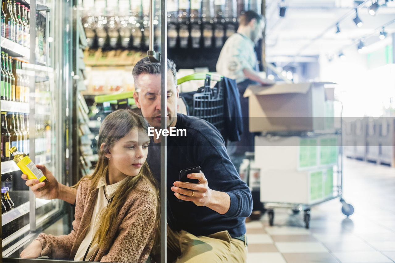 Father and daughter looking at mobile phone while crouching by refrigerator in store