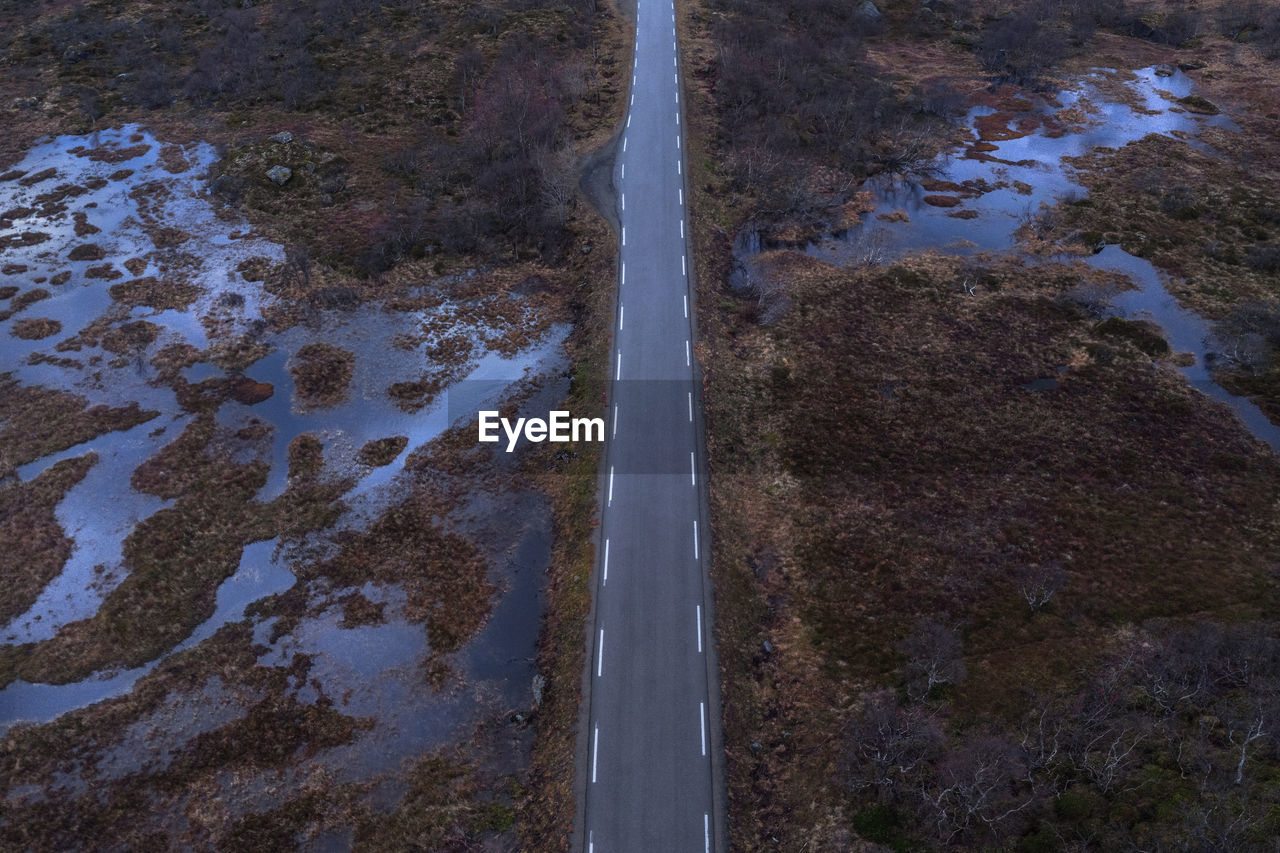 Drone view of long narrow asphalt roadway with making lines running between wetland with withered grass in nature of norway