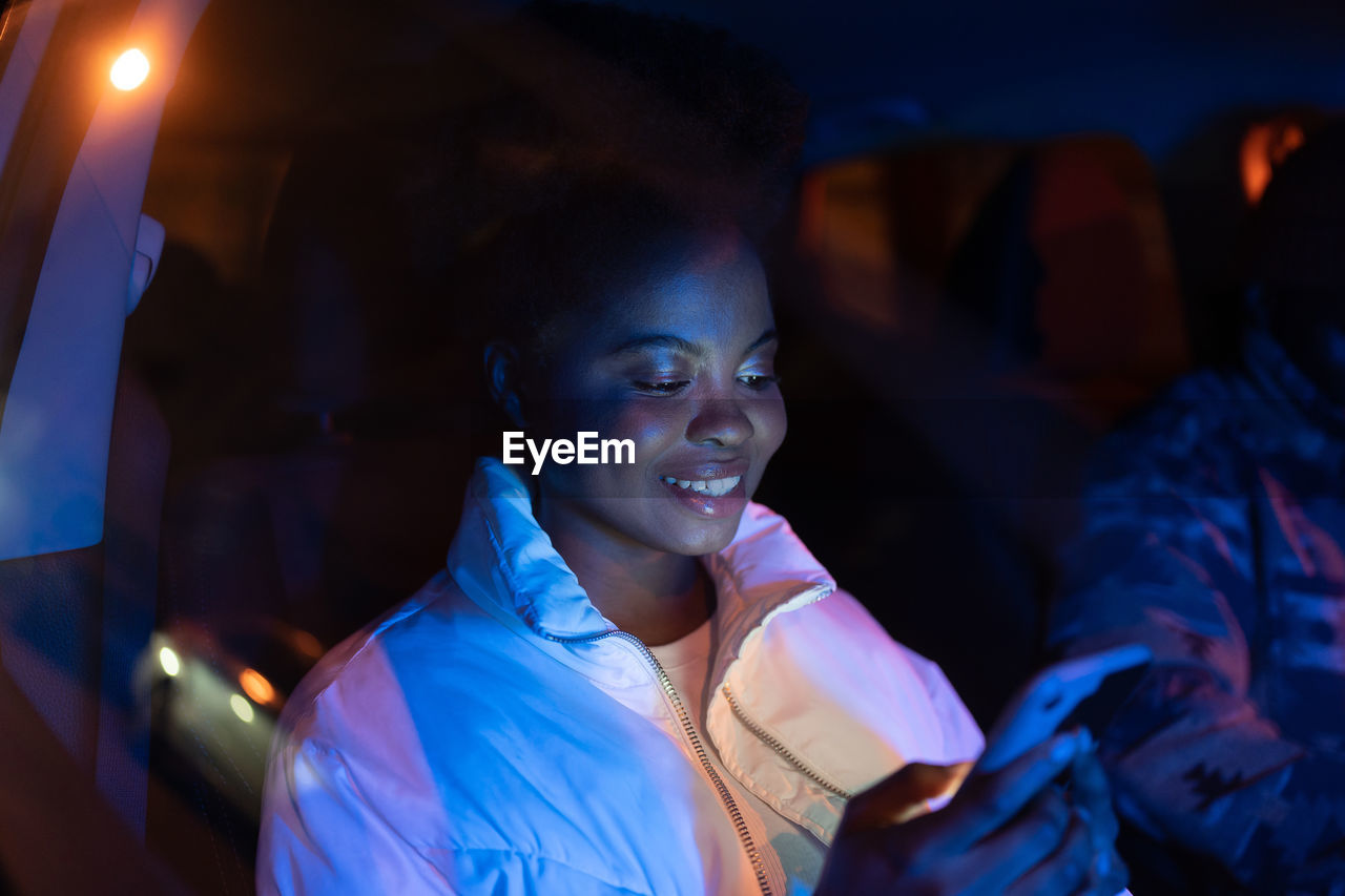 Smiling afro american woman sitting in car on passenger seat with smartphone, using social media app