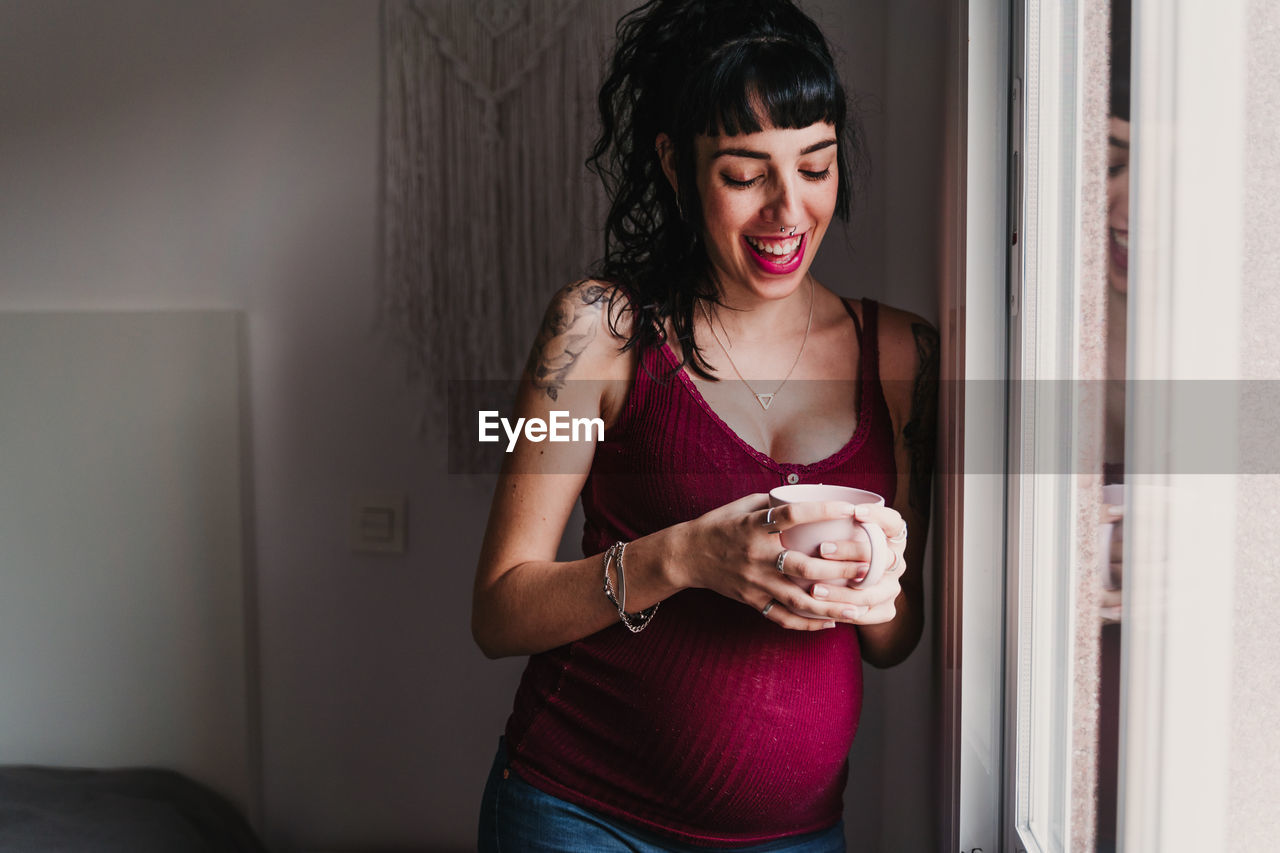 Smiling pregnant woman with mug standing by window at home