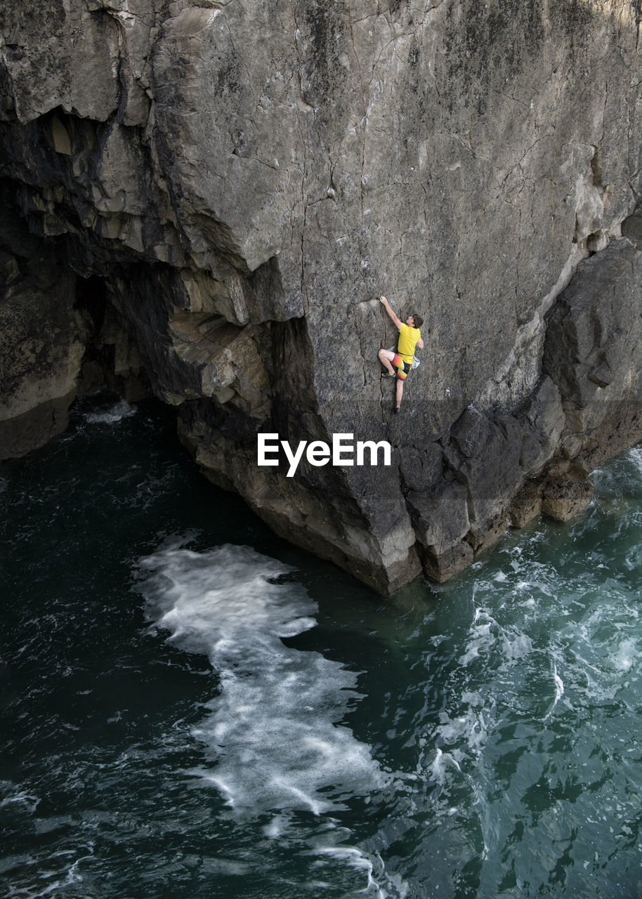 Young male rock climber climbing cliff above water