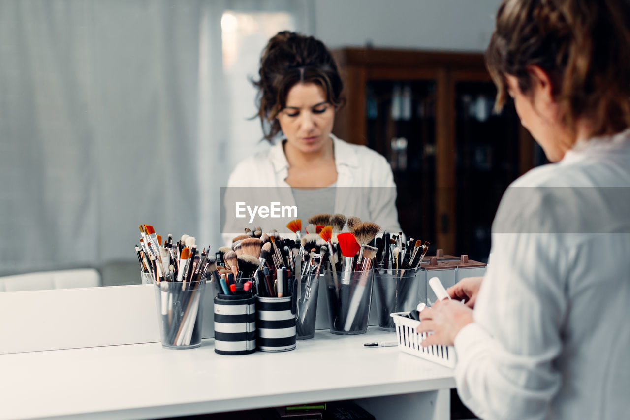 Female visagiste reflecting in mirror standing at table with various makeup supplies in modern beauty studio