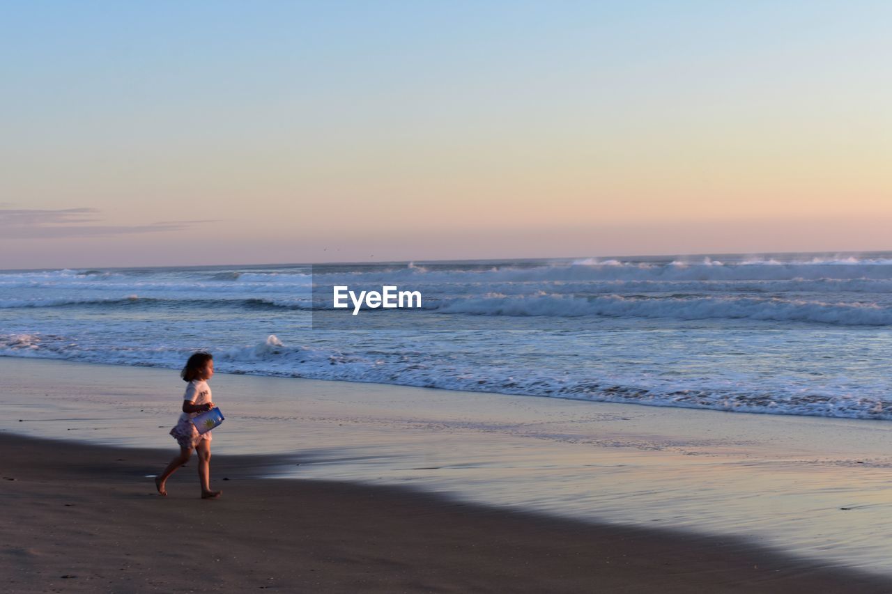 rear view of woman walking at beach against clear sky during sunset