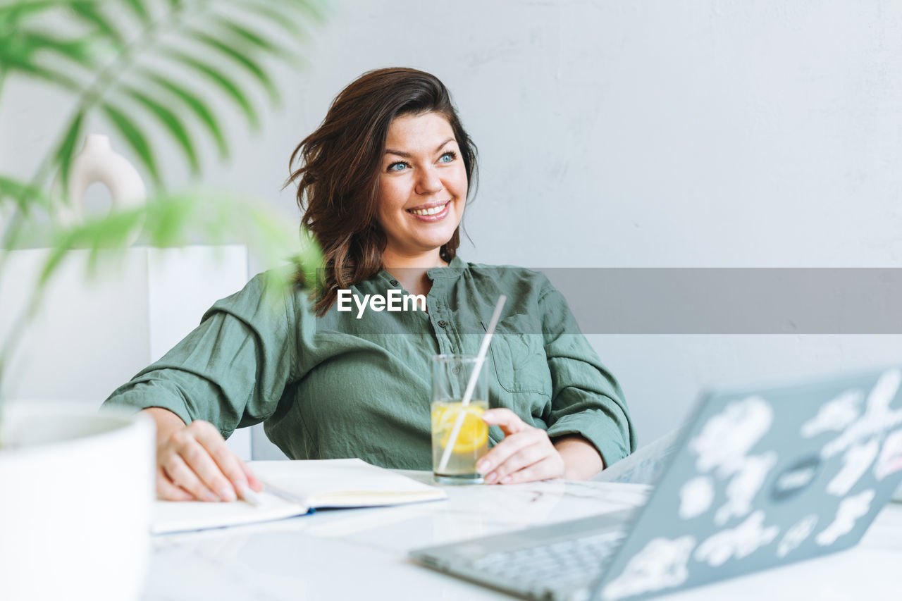 Young smiling brunette woman plus size in casual shirt with lemon water working at laptop in office