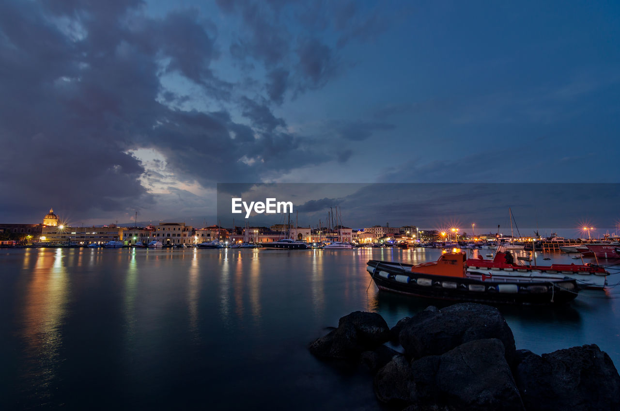 Sea by illuminated city against sky at sunset