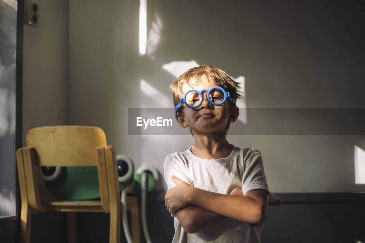 Portrait of boy wearing blue eyeglasses and standing with arms crossed in classroom at kindergarten