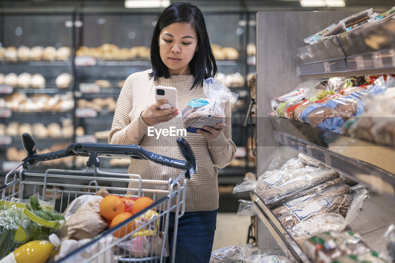 Young woman using smart phone while holding bread package and shopping in supermarket