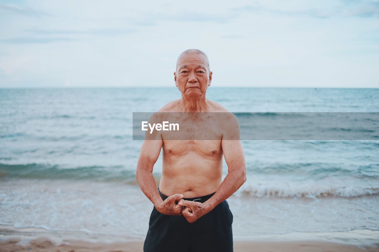 Portrait of shirtless senior man flexing muscles while standing at beach