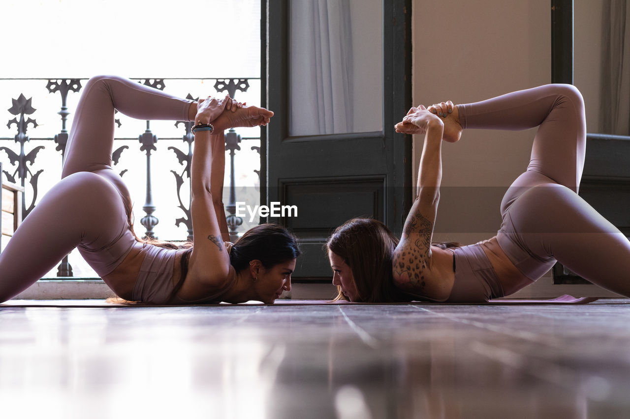 Side view of graceful young hispanic female friends lying on floor and doing stretching exercise during yoga practice together at home