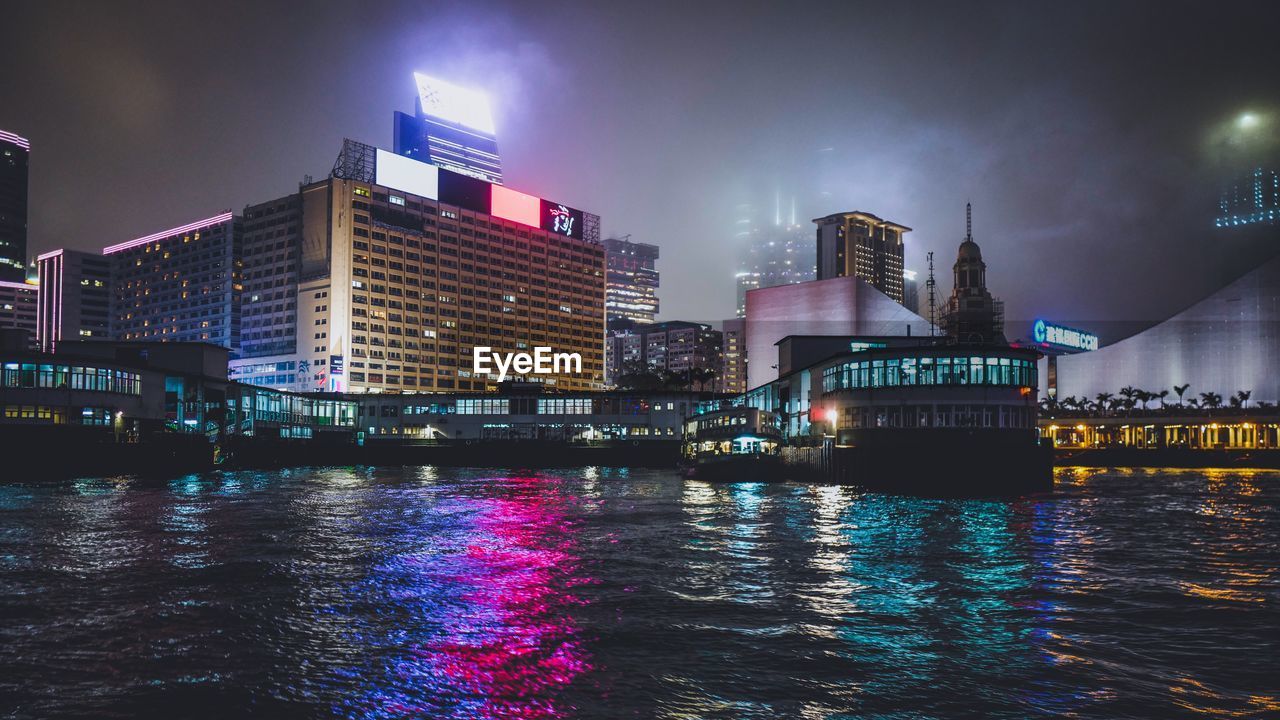 Illuminated buildings in city by sea against sky