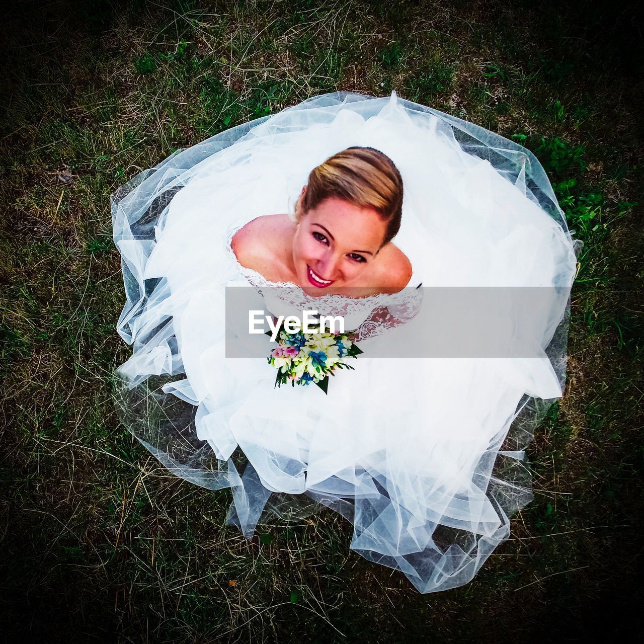 High angle portrait of smiling bride with bouquet sitting on field
