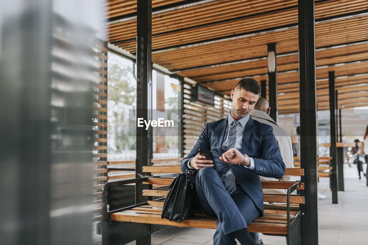 Businessman checking time while sitting on bench at bus stop