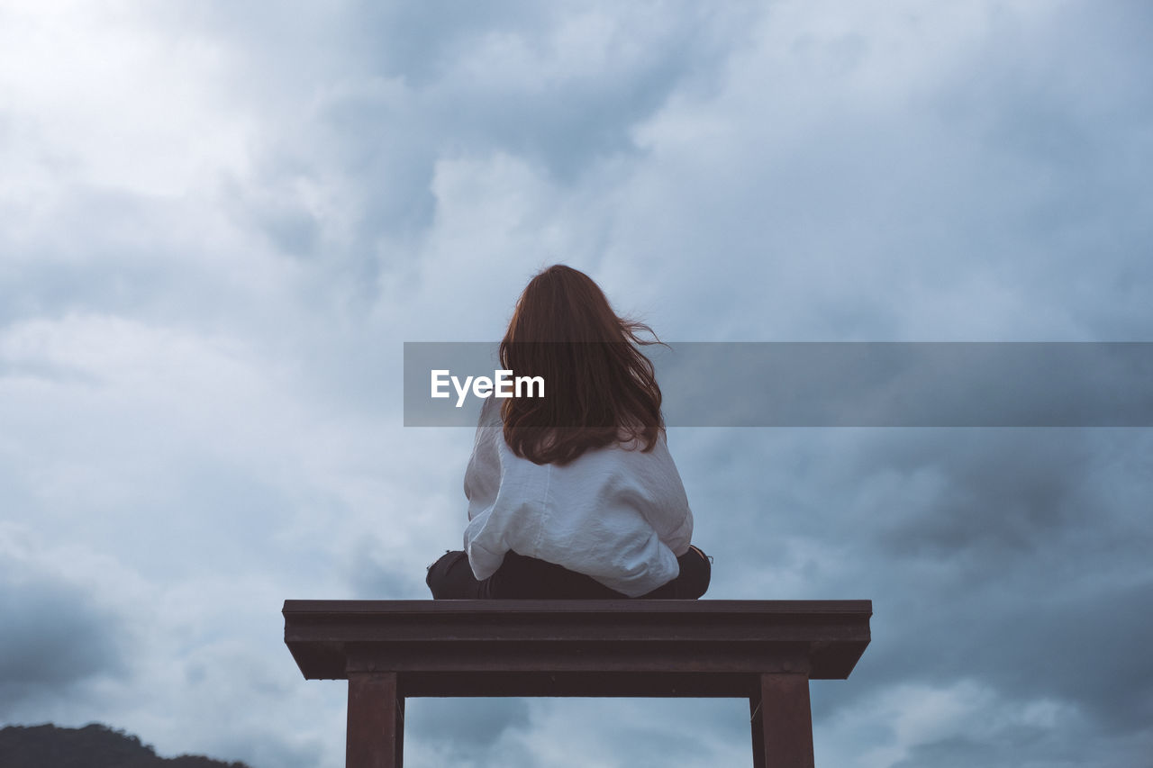 Rear view of woman sitting on bench against cloudy sky