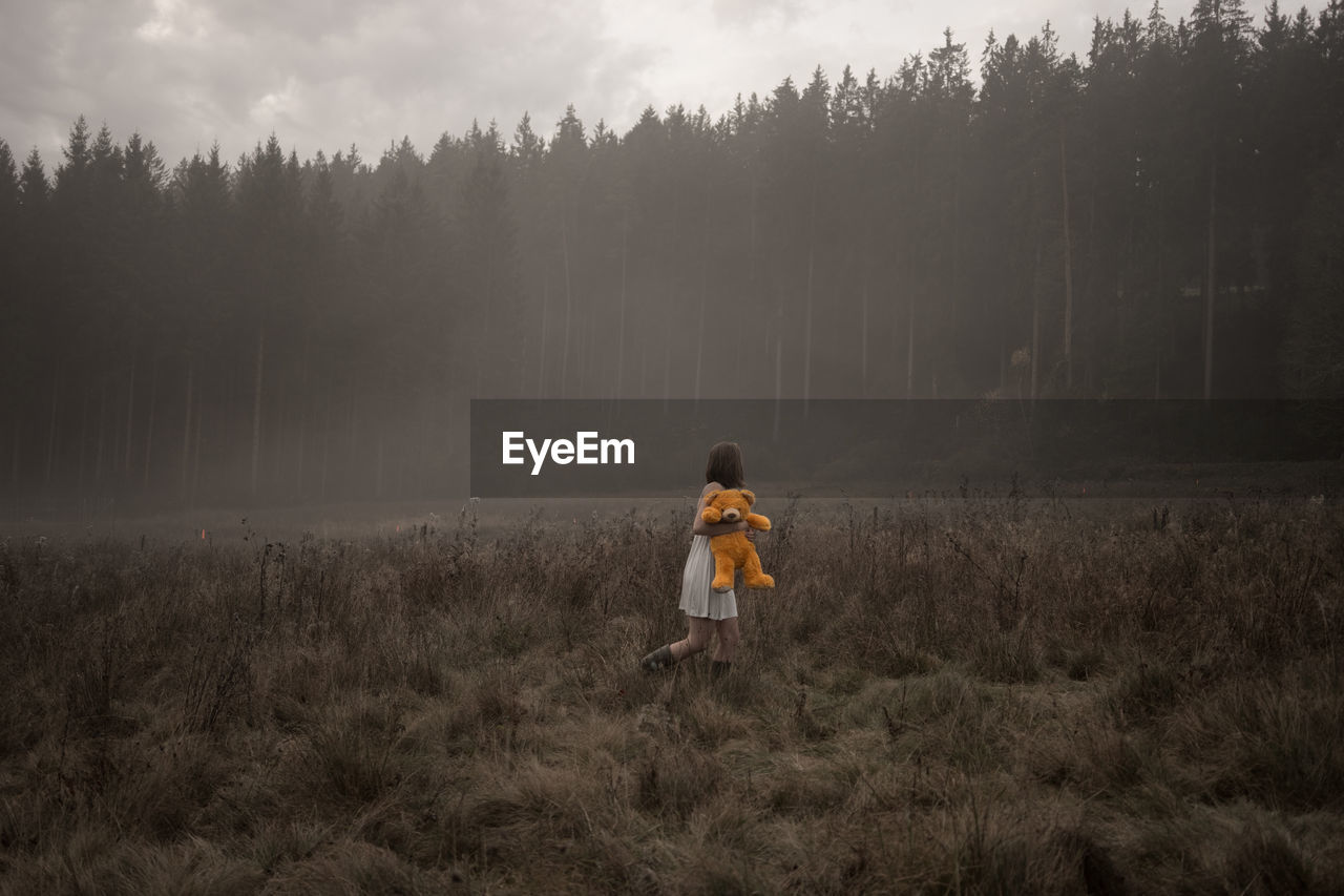 Side view of woman with teddy bear walking on grassy field against trees in forest